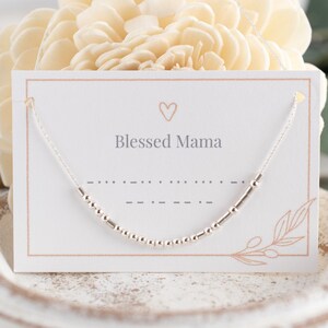 May include: A white card with the text "Blessed Mama" and a small heart above the text. A silver beaded necklace is displayed on the card.