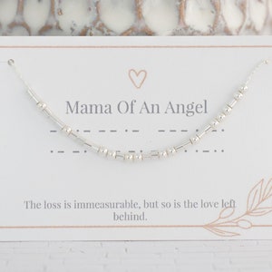 May include: A white card with the text "Mama Of An Angel" and a silver beaded necklace on top. The card also has the text "The loss is immeasurable, but so is the love left behind."