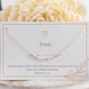 Puede incluir: Un collar de plata con un pequeño dije en forma de corazón y la palabra "Trust" en una tarjeta blanca. La tarjeta también incluye el versículo bíblico Proverbios 3:5, "Confía en el Señor con todo tu corazón, y no te apoyes en tu propia prudencia."