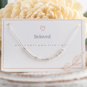 Puede incluir: Un collar de plata con una cadena delicada y pequeñas cuentas. El collar se muestra en una tarjeta blanca con la palabra "Beloved" y un símbolo de corazón.