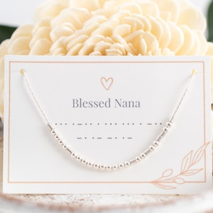May include: A silver necklace with small beads and a heart charm. The necklace is displayed on a white card with the text "Blessed Nana" and a line of braille.