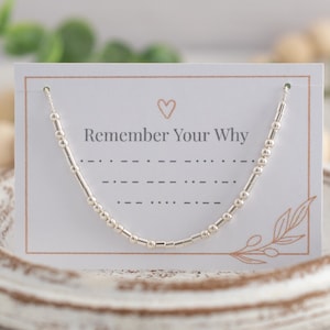 May include: A silver beaded necklace with the words "Remember Your Why" on a white card with a small heart and a branch design.