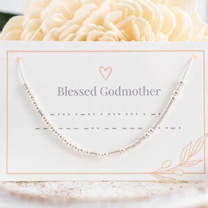 May include: A silver beaded bracelet with the text "Blessed Godmother" on a white card with a small heart.