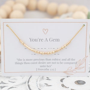May include: A gold necklace with small beads that spells out "You're A Gem" on a white card with the quote "She is more precious than rubies: and all the things thou canst desire are not to be compared unto her [Proverbs 3:15]"