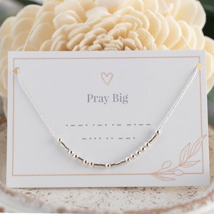 Puede incluir: Un collar de plata con una cadena delicada y pequeñas cuentas. El collar se muestra en una tarjeta blanca con el texto "Pray Big" impreso en ella.