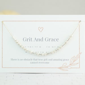 May include: A silver beaded necklace with the words "Grit And Grace" and the quote "There is no obstacle that true grit and amazing grace cannot overcome." on a white card with a pink heart.