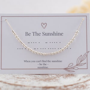 Könnte beinhalten: Ein silbernes Perlenarmband mit dem Text "Be The Sunshine" und einer Braille-Botschaft auf einer weißen Karte mit braunem Rand. Die Karte enthält auch den Text "When you can't find the sunshine - be the - sunshine."