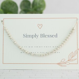 May include: A silver beaded necklace with the words "Simply Blessed" on a white card with a small heart drawn on it.