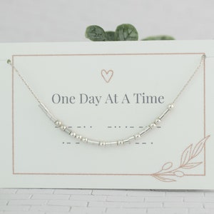 May include: A white card with a silver necklace on it. The necklace has small silver beads and a heart charm. The card says "One Day At A Time" in black text.