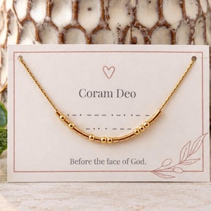 Può includere: Una collana a catena color oro con piccole perline metalliche disposte in un motivo a codice Morse, esposta su un cartoncino rettangolare bianco. Il cartoncino include un'icona a forma di cuore, il testo Coram Deo e la frase Before the face of God.