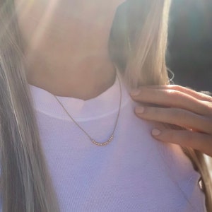 May include: A close-up of a person wearing a white long-sleeved shirt with a delicate gold necklace featuring a row of small beads.