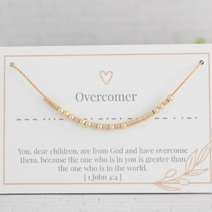 Pode incluir: Pulseira de contas douradas com um cartão que diz "Overcomer" e um versículo bíblico de 1 João 4:4. O cartão tem um pequeno coração e um desenho floral.