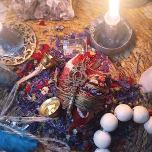 May include: A lit candle in a metal holder is the focal point, surrounded by spiritual items. These include a key, bells, crystals, dried flowers, and white beads. The scene is set on a woven mat, creating a mystical aesthetic. A blue feather is also visible.