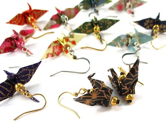 May include: A collection of origami crane earrings with various patterns and colors, including gold, black, blue, red, and white. The earrings are made with paper and have gold bead accents.