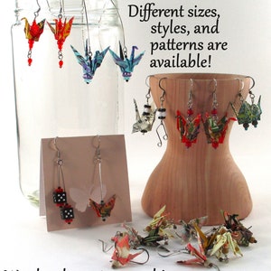 Op de afbeelding: Een assortiment origami-kraanvogel oorbellen in verschillende kleuren en patronen. De oorbellen worden getoond op een houten standaard en in een glazen pot. De tekst "Different sizes, styles, and patterns are available!" staat boven de oorbellen. De tekst "We also do custom work! Check us out at charmcityfab.store!" staat onder de oorbellen.