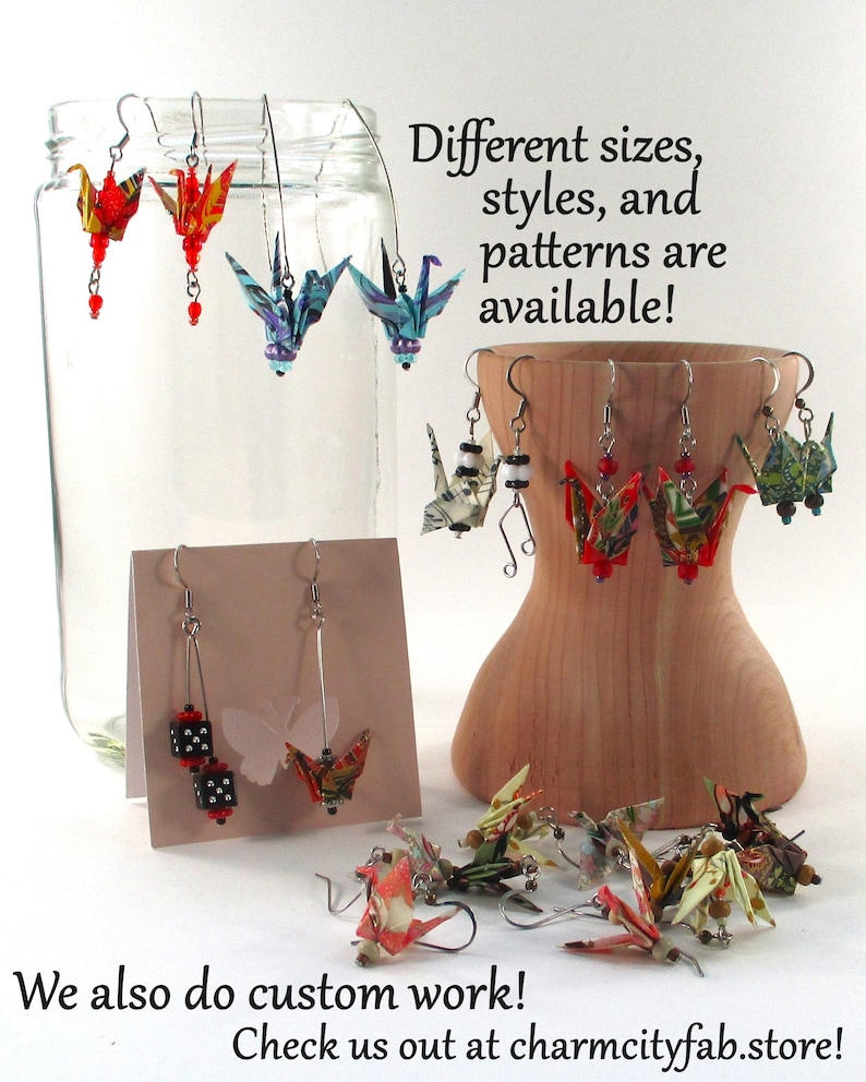 Op de afbeelding: Een assortiment origami-kraanvogel oorbellen in verschillende kleuren en patronen. De oorbellen worden getoond op een houten standaard en in een glazen pot. De tekst "Different sizes, styles, and patterns are available!" staat boven de oorbellen. De tekst "We also do custom work! Check us out at charmcityfab.store!" staat onder de oorbellen.