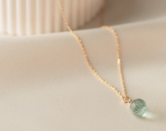 Puede incluir: Un delicado collar de cadena dorada con un solo colgante de piedra preciosa redonda de color verde claro. El collar se muestra en una caja de cerámica blanca con tapa.