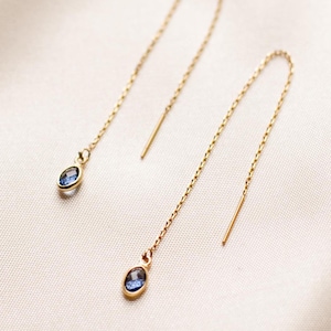 Puede incluir: Un par de pendientes colgantes dorados. Cada pendiente presenta una fina cadena con una pequeña gema azul de forma ovalada en la parte inferior. Los pendientes se muestran sobre un fondo de tela blanquecina.