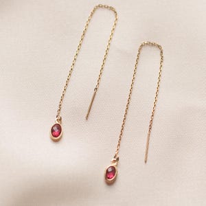 May include: A pair of gold-colored threader earrings. Each earring features a delicate chain with a small, oval-shaped, red gemstone at the bottom. The earrings are displayed against a soft, off-white background.
