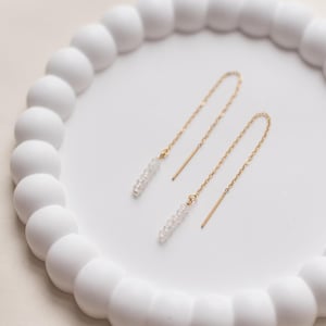 Könnte beinhalten: Ein Paar zarte goldene Kettenohrringe mit kleinen weißen Perlen, die an den Enden hängen. Die Ohrringe werden auf einer weißen runden Schale mit strukturierter Oberfläche präsentiert.