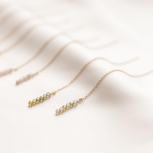 May include: A set of delicate gold chain earrings with small green and yellow gemstone beads. The earrings are displayed on a white fabric background.