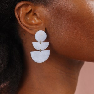 May include: White geometric earrings with three tiers. The top tier is a circle, the middle tier is a half-circle, and the bottom tier is a larger half-circle. The earrings are made of a smooth, white material and have gold accents.