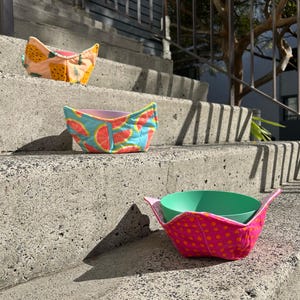 May include: Three fabric bowl cozies with fruit patterns, including pineapples, watermelons, and polka dots. The cozies are displayed on concrete steps, highlighting their colourful designs and use for holding bowls.