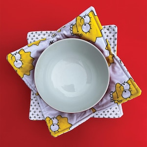 May include: A white ceramic bowl with a yellow and white patterned bowl cosy featuring a cartoon character. The bowl cosy is made of fabric and has a quilted design.