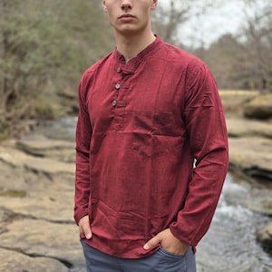 May include: A long-sleeved, burgundy shirt with a mandarin collar and a single chest pocket. The shirt has three wooden buttons and is paired with gray pants. The model is standing outdoors near a stream.