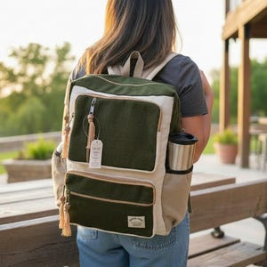 May include: A two-tone backpack featuring a dark olive green main body with cream-colored side panels and trim. The bag includes two front zippered pockets with light-colored tassel pulls, a side pocket holding a silver tumbler, and a small label.
