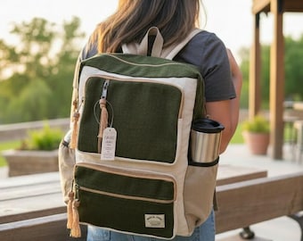 Può includere: Uno zaino bicolore con corpo principale verde oliva scuro e pannelli laterali e finiture color crema. La borsa presenta due tasche frontali con cerniera e tiretti a nappa chiari, una tasca laterale con un bicchiere e una piccola etichetta.