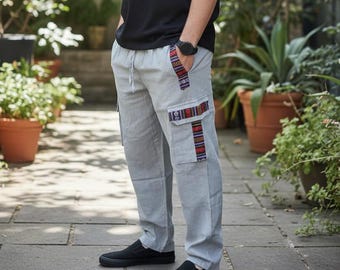 Peut inclure: Une personne porte un t-shirt noir à col rond, un pantalon cargo gris clair avec des motifs décoratifs et des chaussures noires à enfiler. Une montre noire est visible. Le cadre est extérieur avec des plantes en pot.