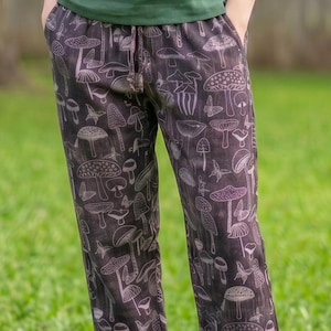 May include: A person wearing a short-sleeved, dark green shirt and dark pants with a mushroom pattern. The pants have a drawstring waist and a variety of mushroom illustrations in white. The person is standing on green grass.
