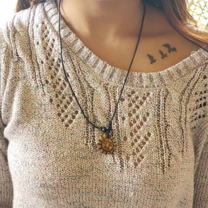 May include: A black cord necklace with a wooden sun pendant. The pendant has a smiling face carved into it. The necklace is worn by a person with a small tattoo of three birds on their collarbone.