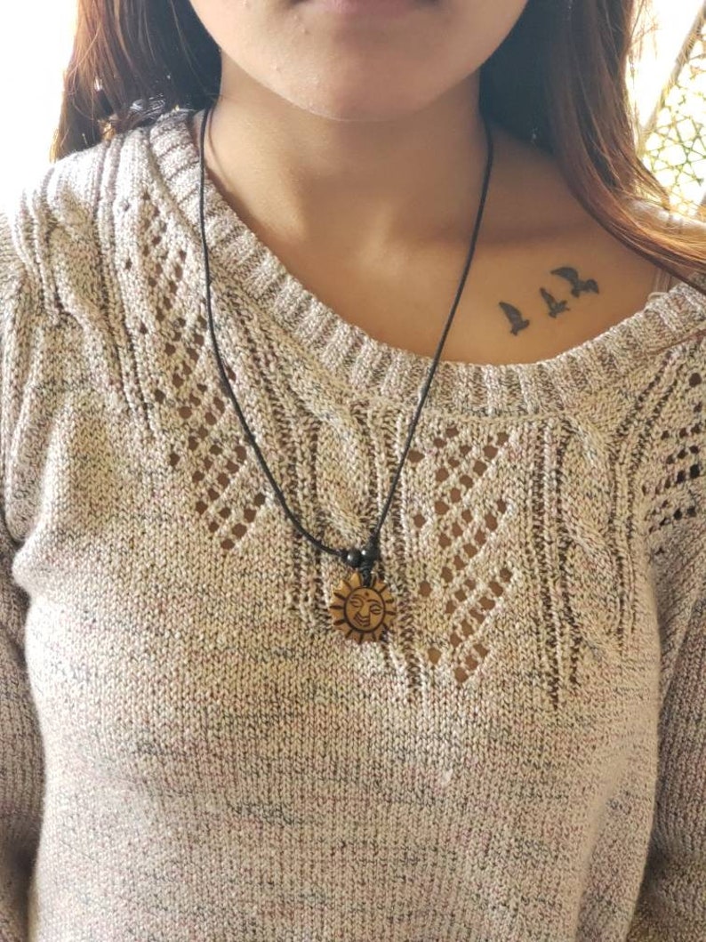 May include: A black cord necklace with a wooden sun pendant. The pendant has a smiling face carved into it. The necklace is worn by a person with a small tattoo of three birds on their collarbone.