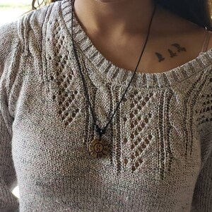 May include: A woman wearing a light brown sweater with a black cord necklace and a round wooden pendant with a yin yang symbol. The pendant is hanging on the woman's chest. The woman has a small tattoo of three birds on her collarbone.