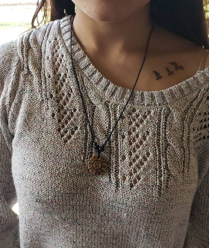 May include: A woman wearing a light brown sweater with a black cord necklace and a round wooden pendant with a yin yang symbol. The pendant is hanging on the woman's chest. The woman has a small tattoo of three birds on her collarbone.
