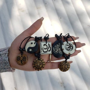 May include: A hand holding six necklaces with different pendants. The pendants are made of wood and feature various designs, including a yin yang symbol, a sun, a face, and the word "Om" in Sanskrit. The necklaces are strung on black cords with black beads.