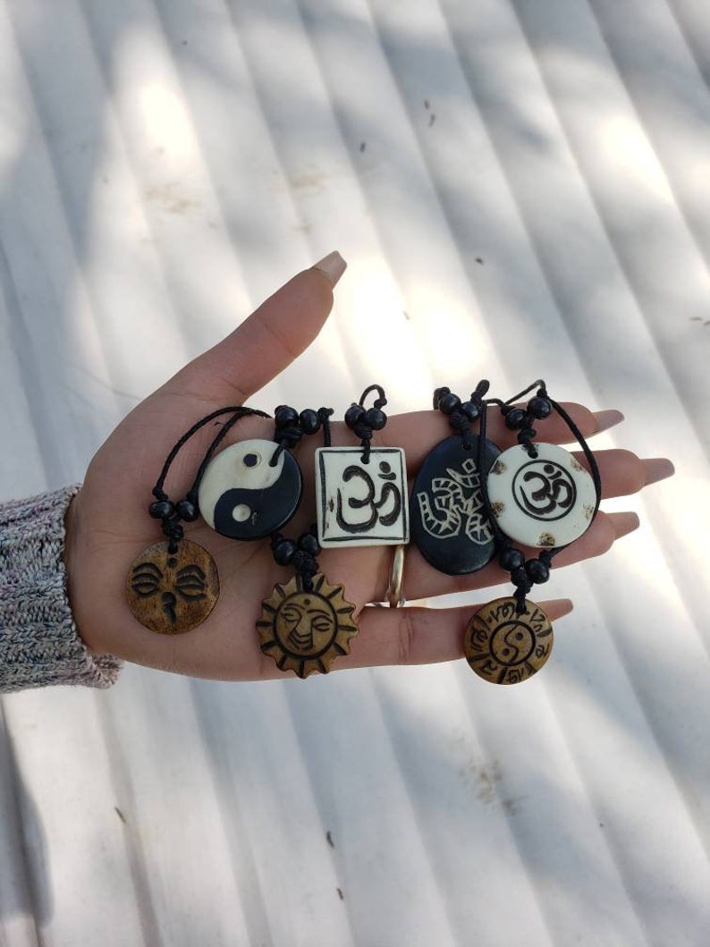 May include: A hand holding six necklaces with different pendants. The pendants are made of wood and feature various designs, including a yin yang symbol, a sun, a face, and the word "Om" in Sanskrit. The necklaces are strung on black cords with black beads.