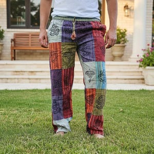 Peut inclure: Pantalon en patchwork présentant un motif multicolore avec divers panneaux de tissu rectangulaires dans des tons de rouge, violet, vert et bleu. Le pantalon est décoré de symboles noirs imprimés, dont le signe Om et des yeux stylisés, et possède un cordon.