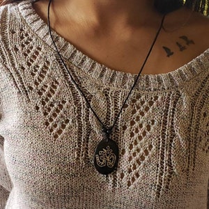 May include: A black necklace with an oval pendant featuring an engraved Om symbol. The necklace is worn by a person with a tattoo of three birds on their shoulder.