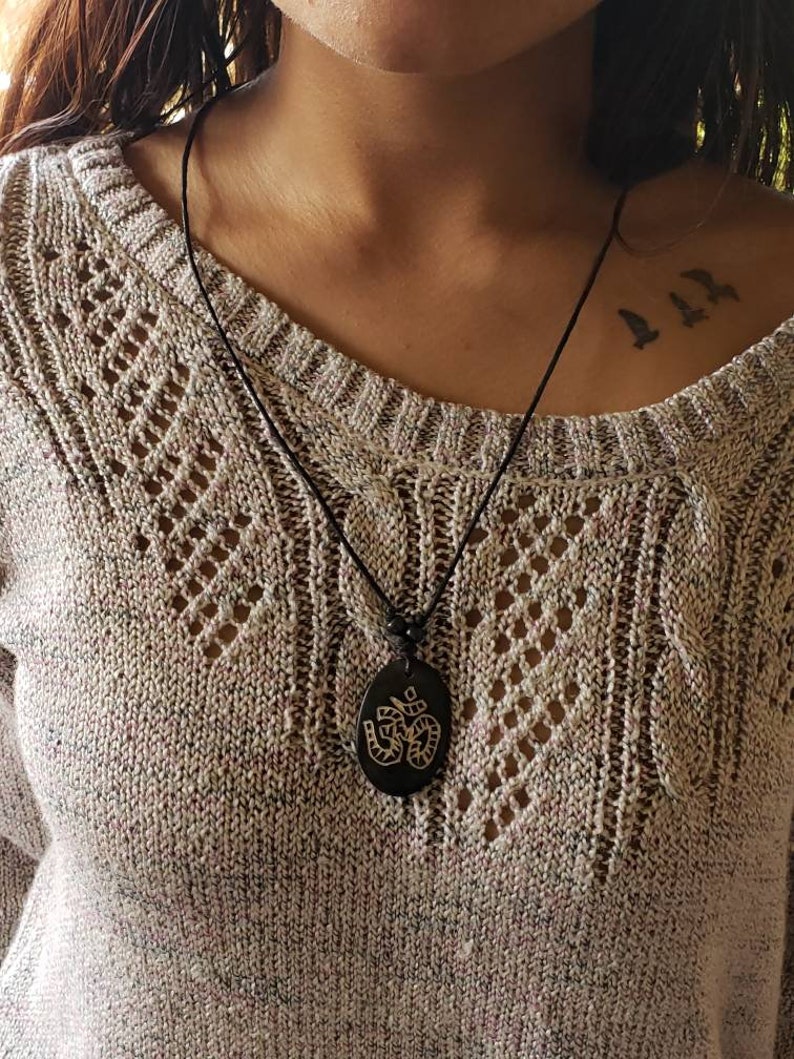 May include: A black necklace with an oval pendant featuring an engraved Om symbol. The necklace is worn by a person with a tattoo of three birds on their shoulder.