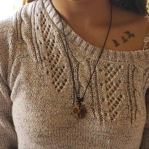 May include: A person wearing a beige sweater with a black cord necklace and a wooden pendant. The pendant has a carved design. The person has a tattoo of three birds on their collarbone.