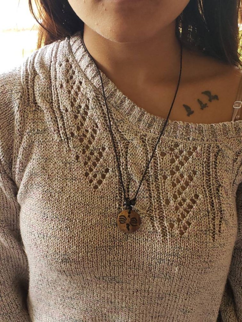 May include: A person wearing a beige sweater with a black cord necklace and a wooden pendant. The pendant has a carved design. The person has a tattoo of three birds on their collarbone.