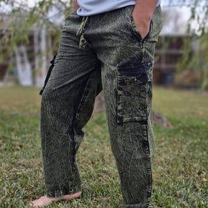 Op de afbeelding: Groene en zwarte cargobroek met een textuurpatroon. De broek heeft zakken aan de zijkanten en een taille met trekkoord. De persoon staat op gras.