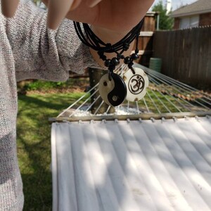 May include: A black cord necklace with two white pendants. One pendant features a yin yang symbol and the other features an Om symbol.