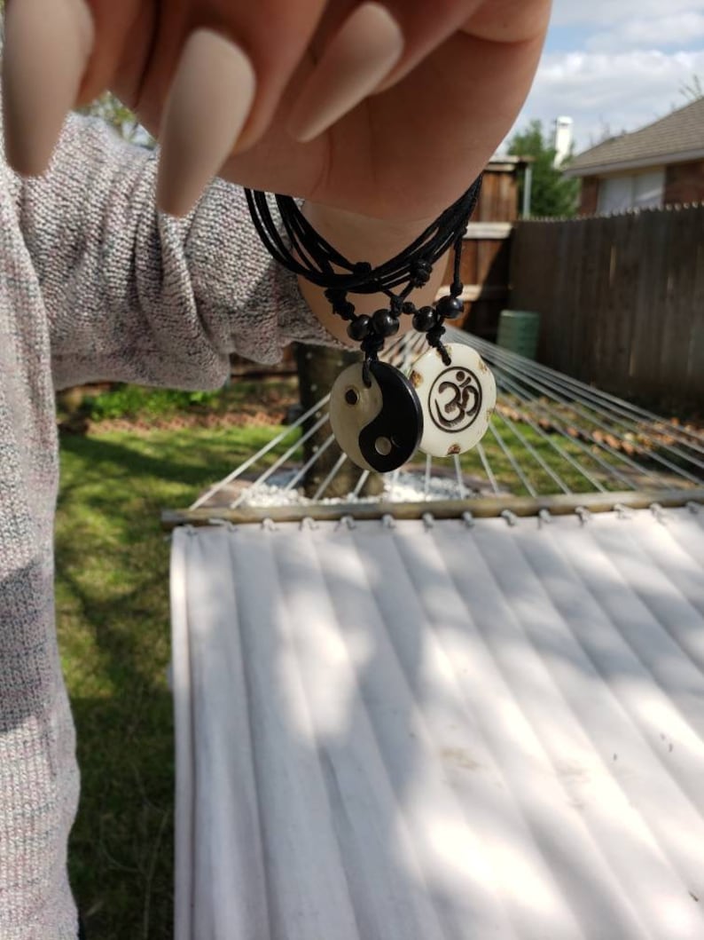 May include: A black cord necklace with two white pendants. One pendant features a yin yang symbol and the other features an Om symbol.