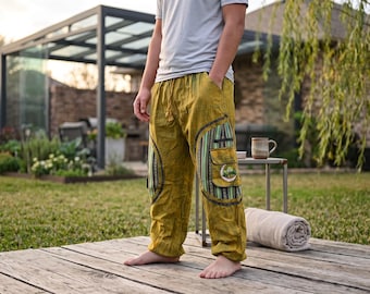 May include: Mustard yellow loose-fitting trousers featuring an elastic drawstring waistband, side pockets, and large cargo pockets on the thighs with vertical green, black, and white striped fabric panels and a circular patch on the left pocket.