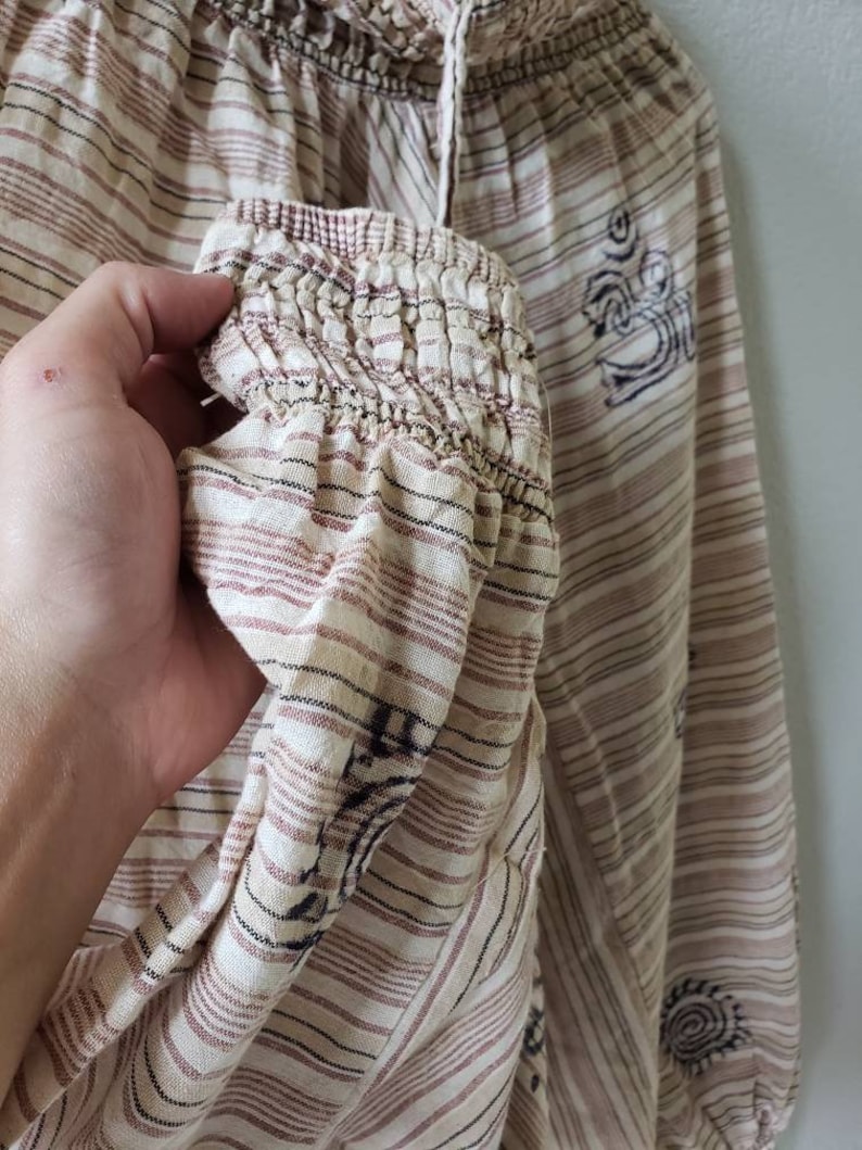 May include: A white and brown striped top with a gathered elastic neckline and long sleeves. The top has a brown and black printed design with a symbol that looks like an Om.