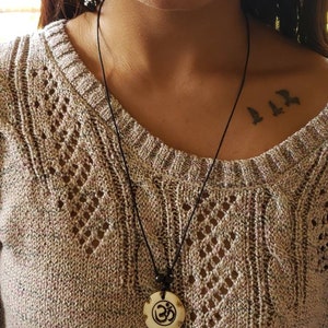 May include: A black cord necklace with a wooden pendant featuring the Om symbol. The pendant is carved with intricate details and has a light brown color.
